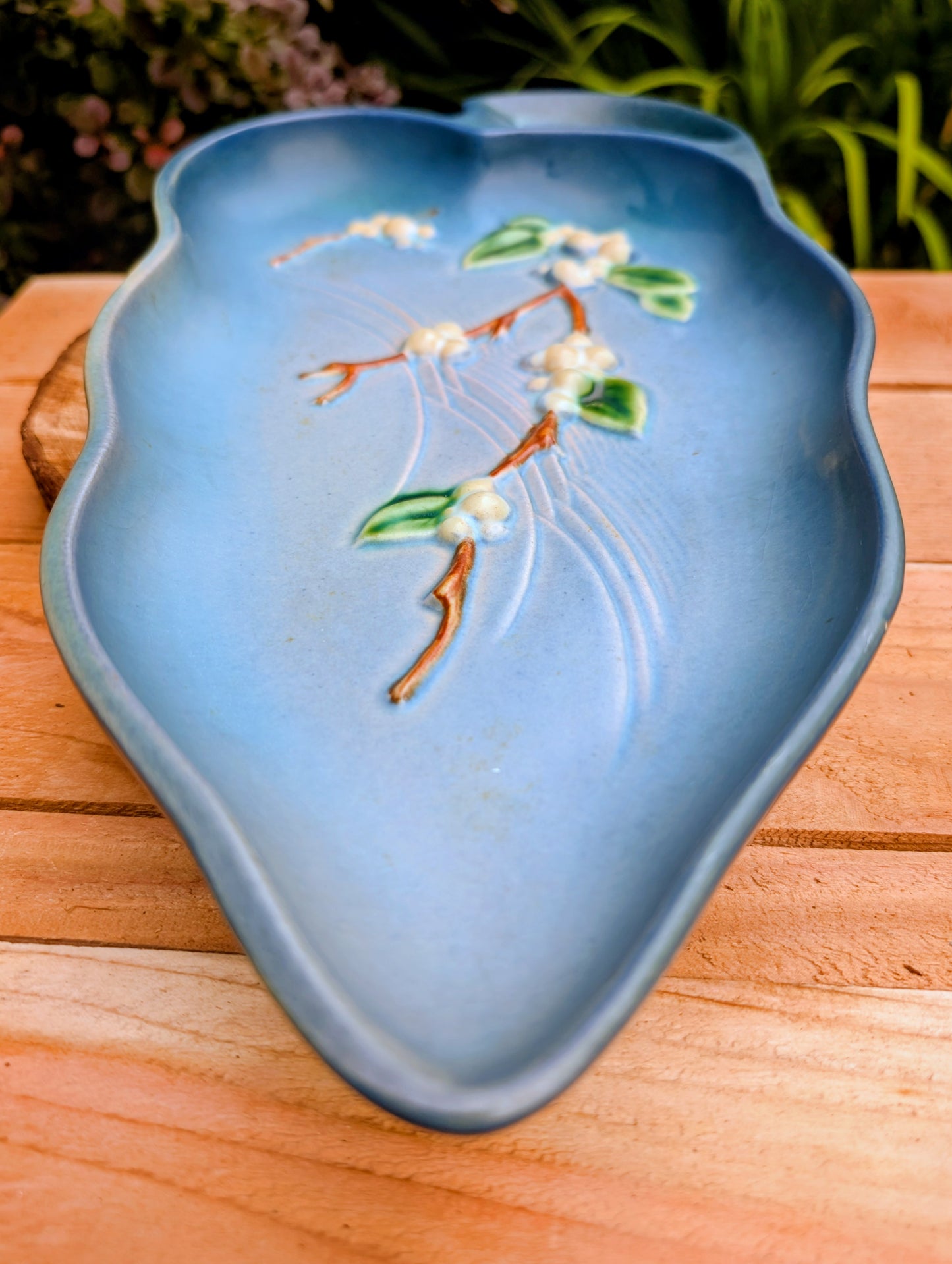 A light blue ceramic dish with a raised floral branch design in green, white, and brown, resting on a wooden surface with a blurred garden background.