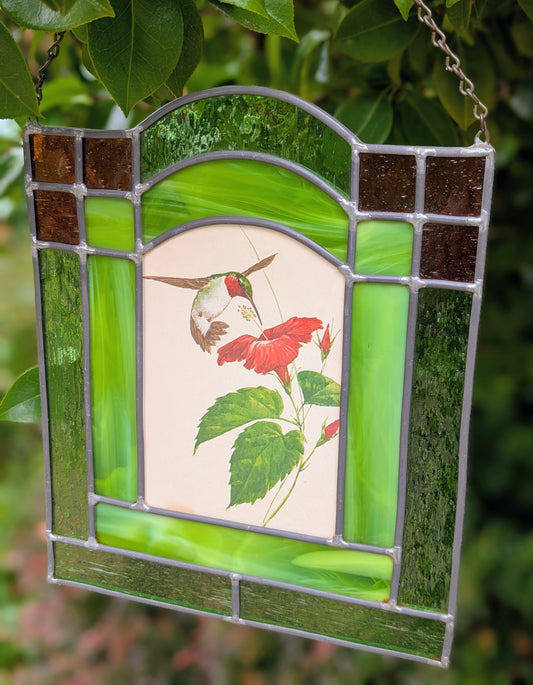 Bring the beauty of a hummingbird in bloom into your home with this handcrafted stained glass hanging panel. Featuring vibrant textured art glass surrounding a detailed hummingbird and hibiscus centerpiece, it catches natural light beautifully and adds year-round color to any window, sunroom, or garden-inspired space.