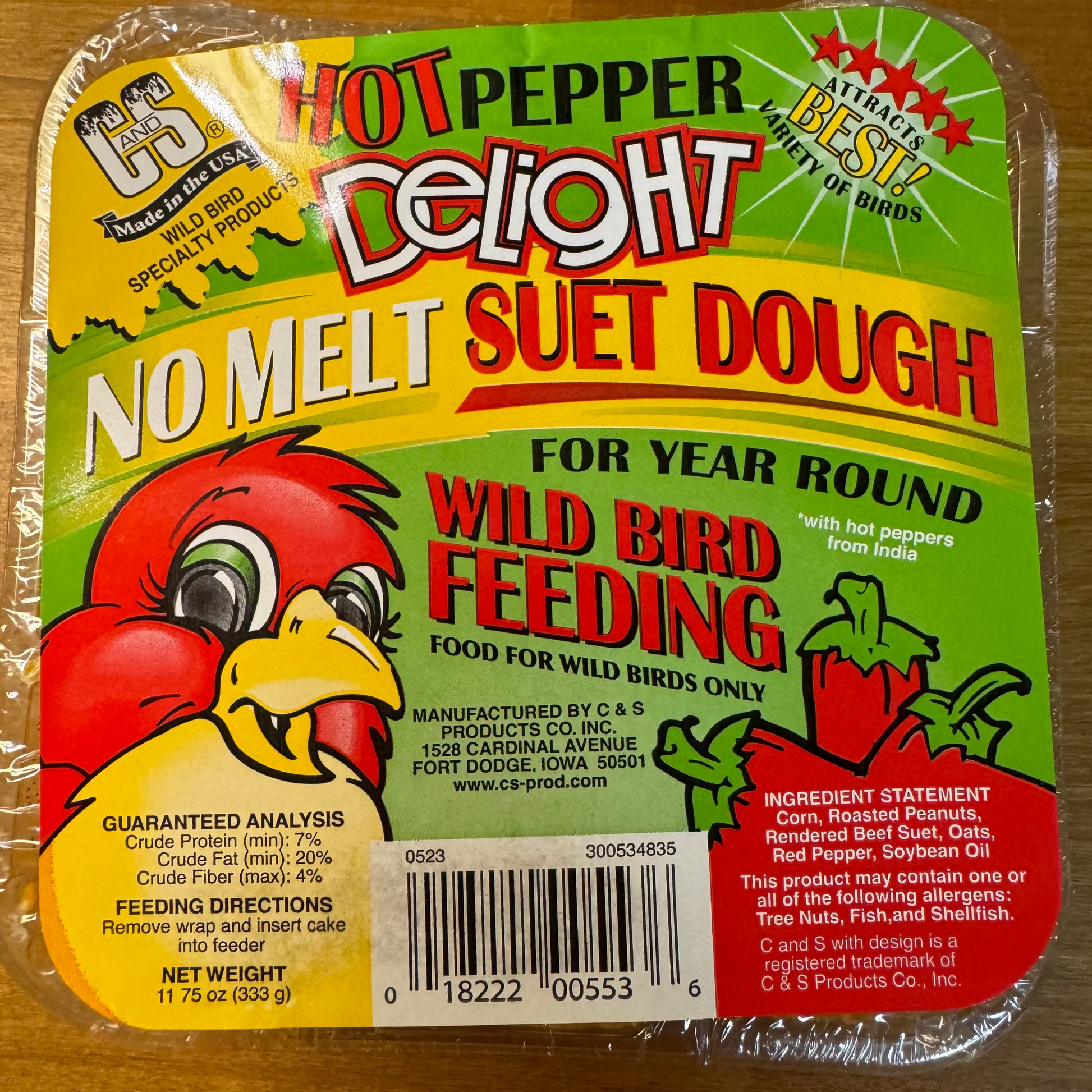 A packaged tray of Hot Pepper Delight No Melt Suet Dough bird feeder on a wooden surface. The packaging is clear plastic wrapping a green, yellow, and red tray with illustrations of a red bird and hot peppers.