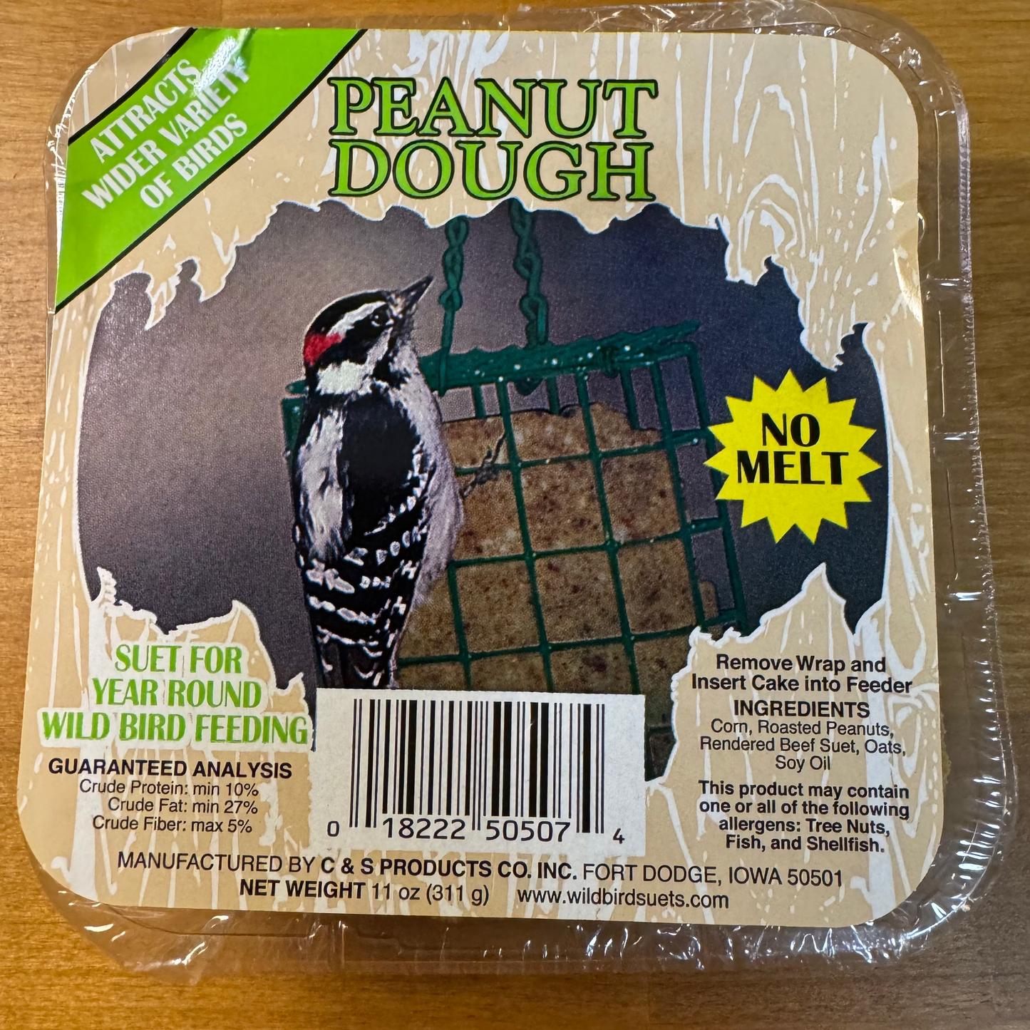 A packaged bird feeder product labeled 'Peanut Dough' on a wooden surface. The square clamshell package contains a nut cake and has a printed front label showing a woodpecker, a green mesh feeder insert, a bright green 'Attacks Wild Variety of Birds' banner, and a yellow 'No Melt' starburst.