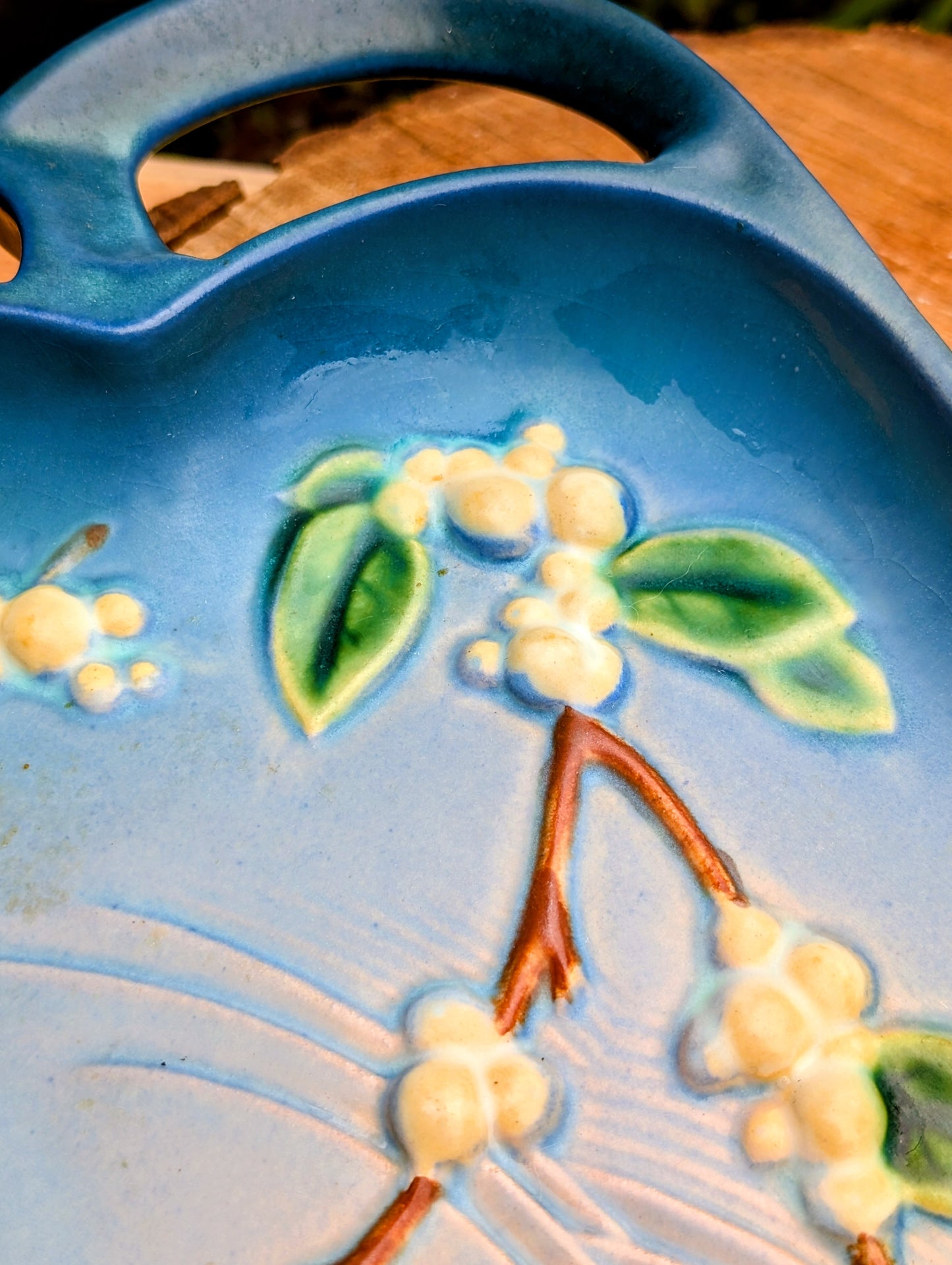 Close-up of a blue ceramic serving tray with embossed floral details of green leaves, cream blossoms, and brown branches, featuring a handle at the top.