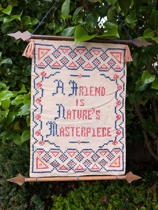 Celebrate the beauty of friendship and handcrafted artistry with this vintage cross-stitch sampler featuring the timeless quote, "A Friend Is Nature's Masterpiece." Mounted on a decorative carved wooden hanger with tassels, this charming textile brings warmth and cottage-inspired character to any home.