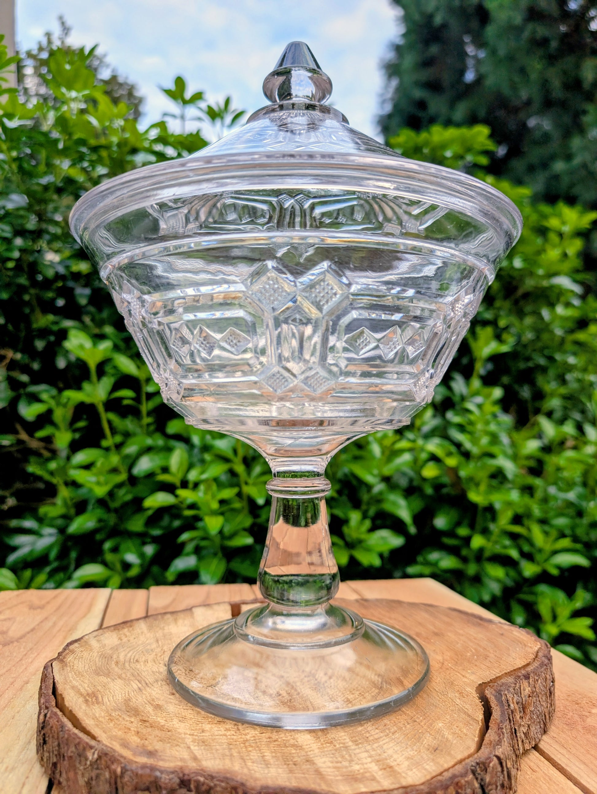 An elegant early 20th-century pressed glass covered compote featuring bold geometric Art Deco styling and a striking pedestal silhouette. Although the pedestal has visible stress cracks, the original lid, impressive scale, and architectural design make it a beautiful decorative display piece.