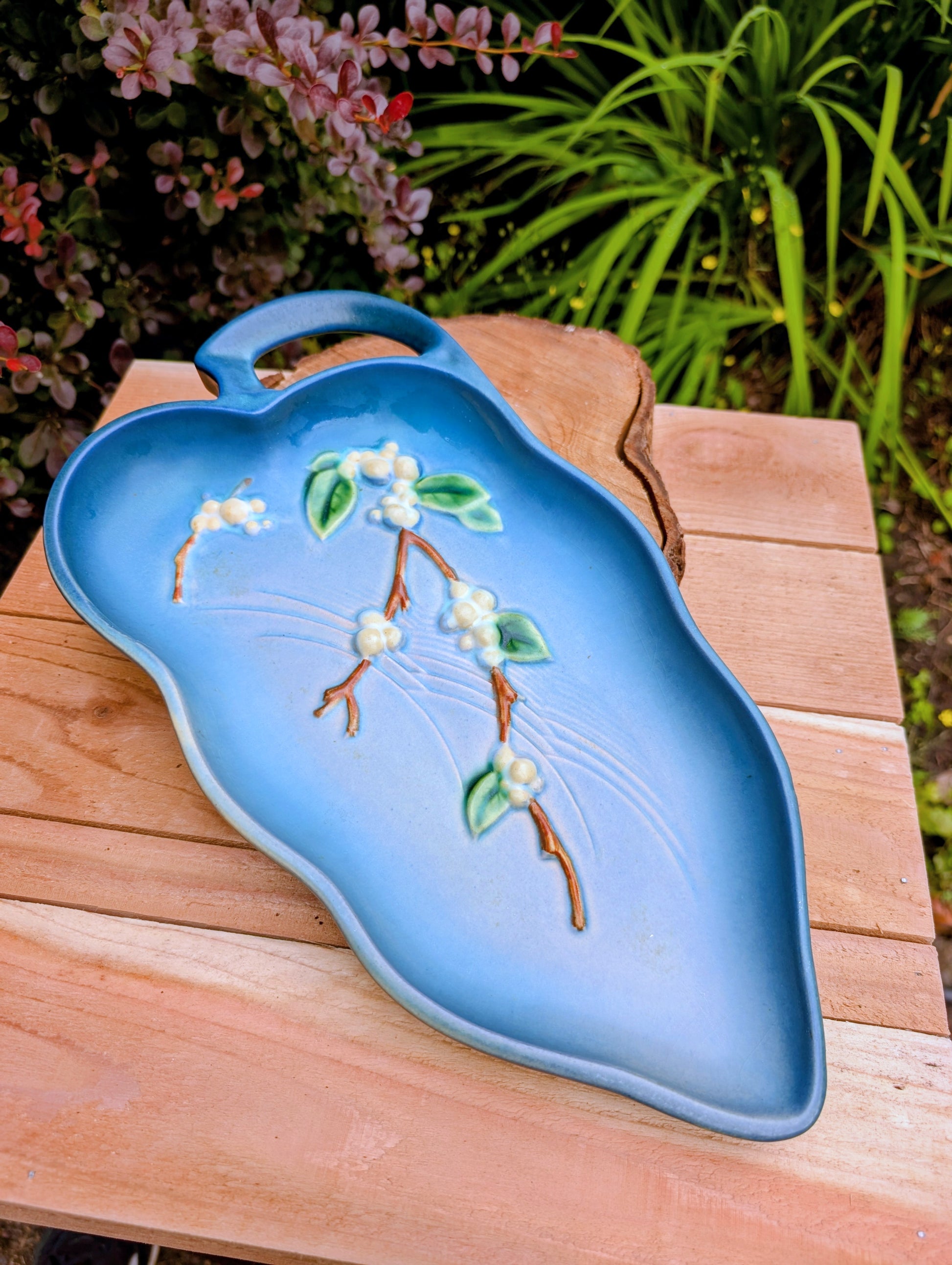 A light blue ceramic serving platter with a handle, decorated with a raised branch motif featuring white blossoms and green leaves, displayed on a wooden table with a garden background.
