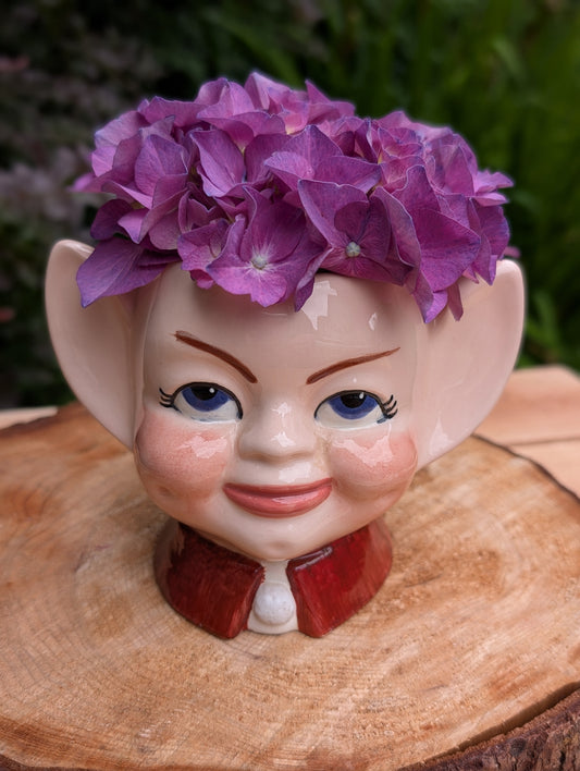 Bring a touch of mid-century whimsy to your home with this vintage ceramic pixie head planter. Hand-painted with expressive blue eyes and oversized pointed ears, this charming planter is perfect for displaying succulents, air plants, or seasonal greenery while adding nostalgic character to any space.