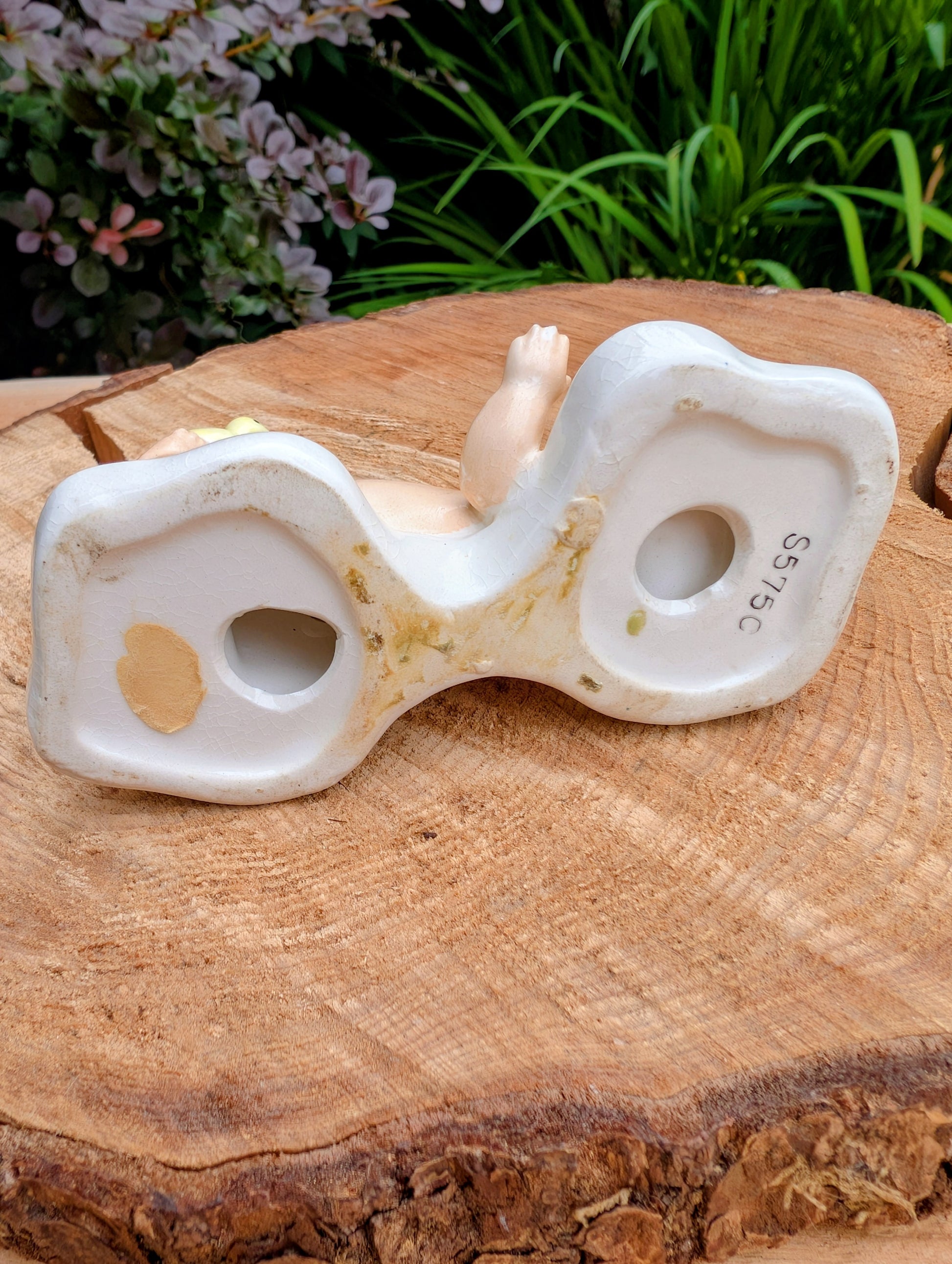 A vintage white ceramic doll's head and upper torso sculpture displayed on a rustic wood slice with green foliage in the background.