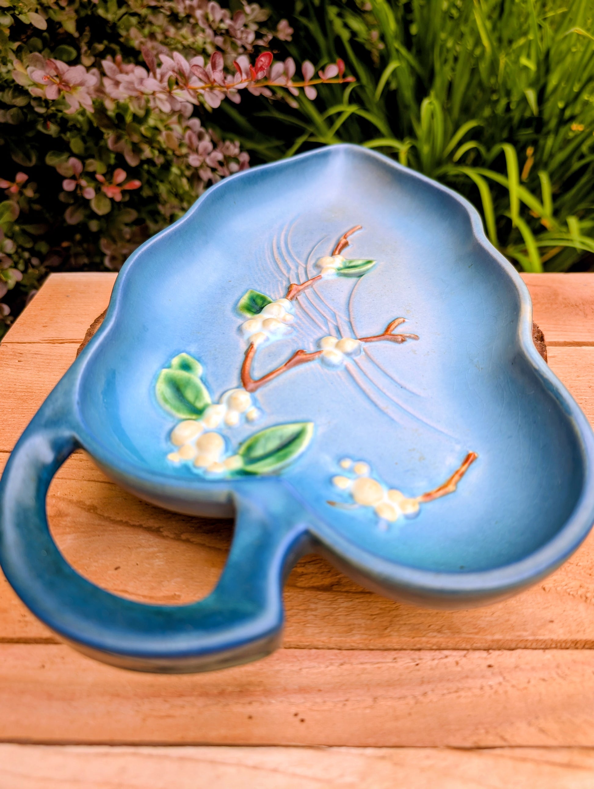 A light blue ceramic serving tray with a handle, decorated with embossed cherry blossom branches and white flowers, resting on a wooden surface with green foliage in the background.