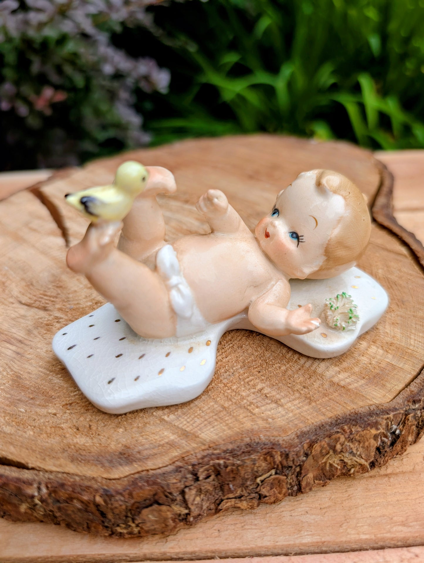 Vintage-style ceramic baby figurine lying on its back on a white patterned mat, holding a small yellow-green duckling, displayed on a rustic wood slice with blurred green foliage in the background.