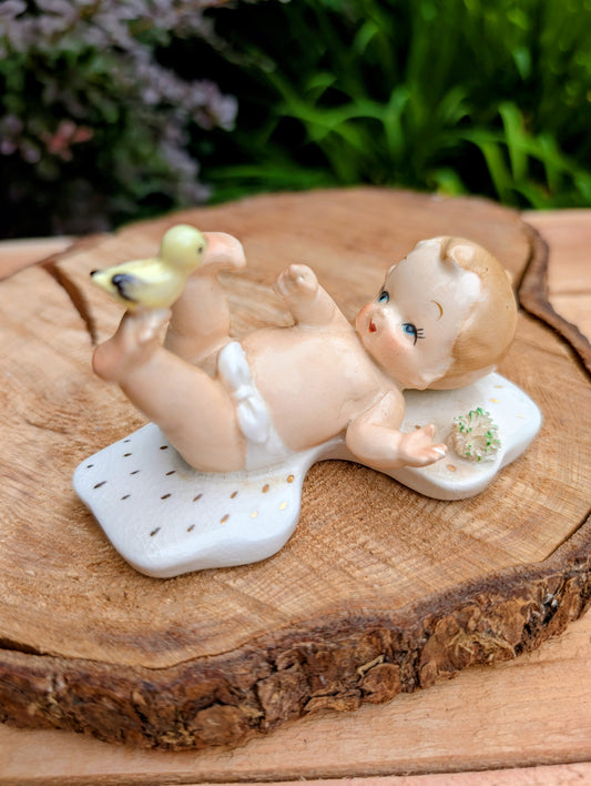 Vintage-style ceramic baby figurine lying on its back on a white patterned mat, holding a small yellow-green duckling, displayed on a rustic wood slice with blurred green foliage in the background.