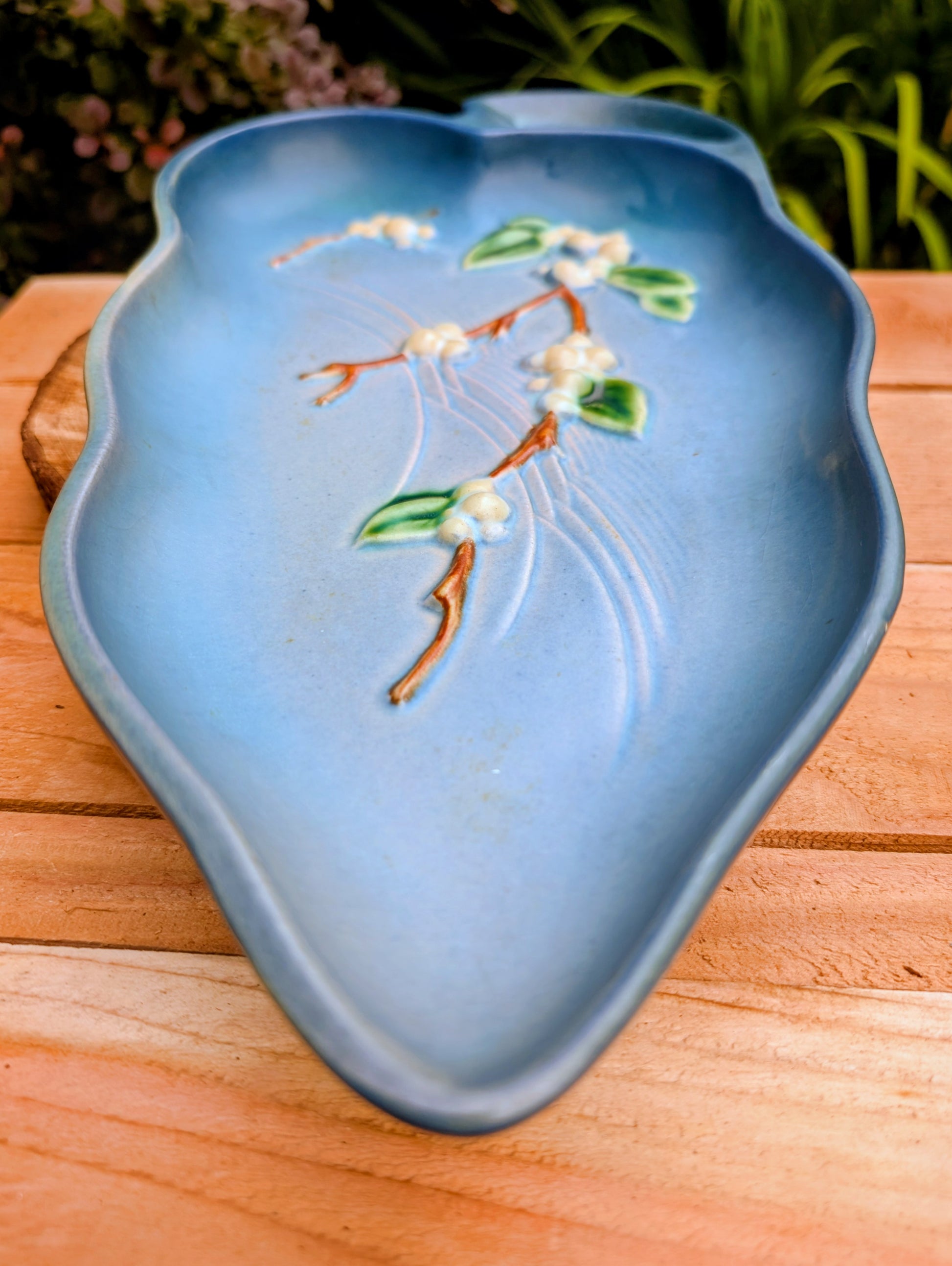 A light blue ceramic dish with a raised floral branch design in green, white, and brown, resting on a wooden surface with a blurred garden background.