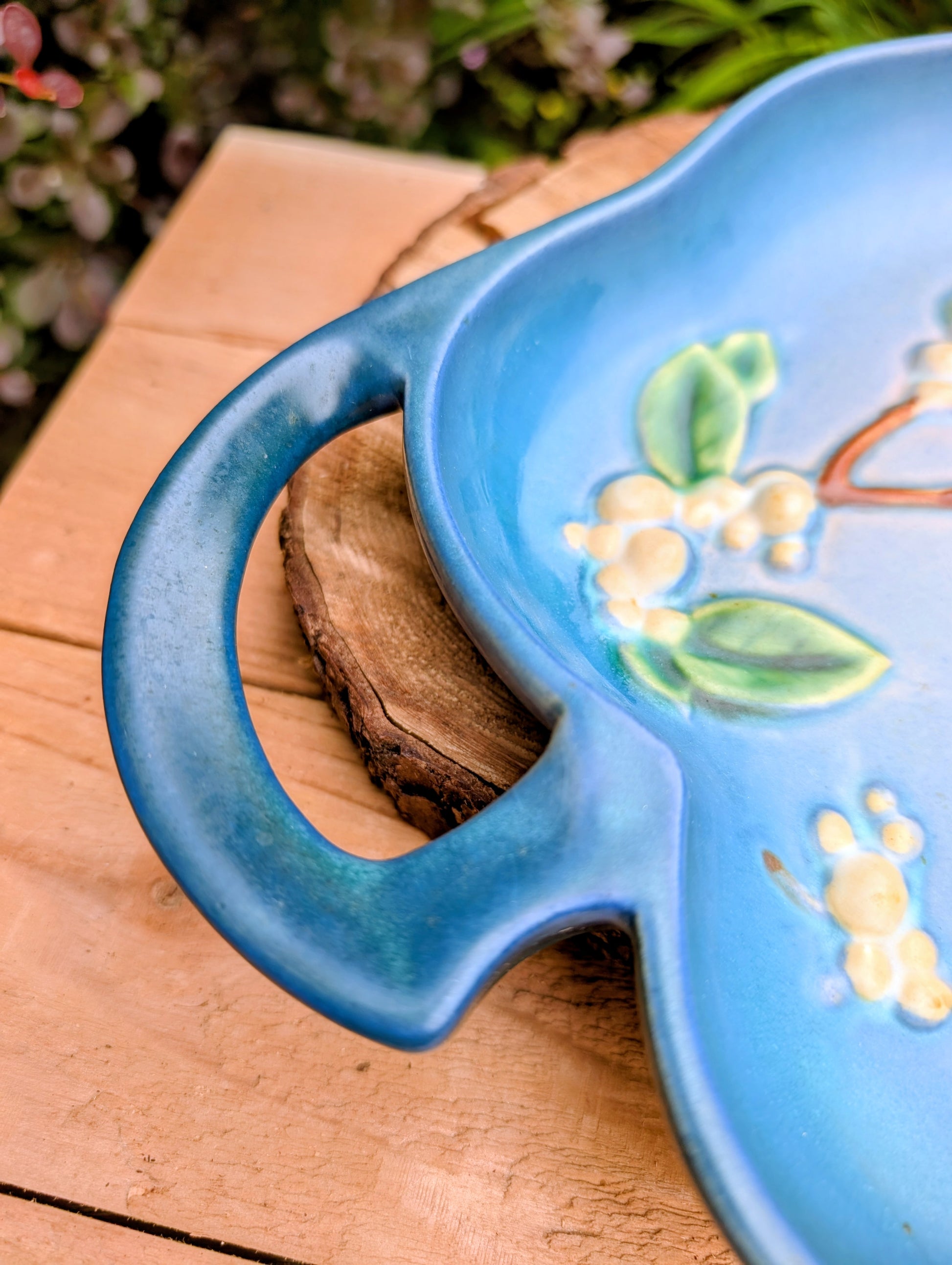 A close-up of a blue ceramic serving platter with a handle, decorated with raised cream flowers and green leaves, resting on a rustic wooden slice over a wooden table.