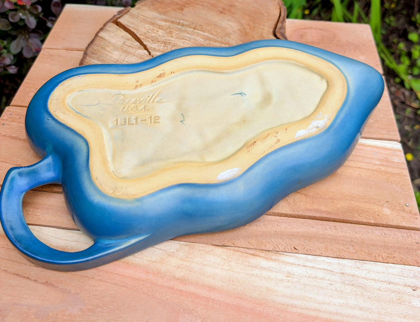 A vintage ceramic planter or decorative dish with a blue glazed exterior and a cream-colored interior, resting on a wooden table outdoors.