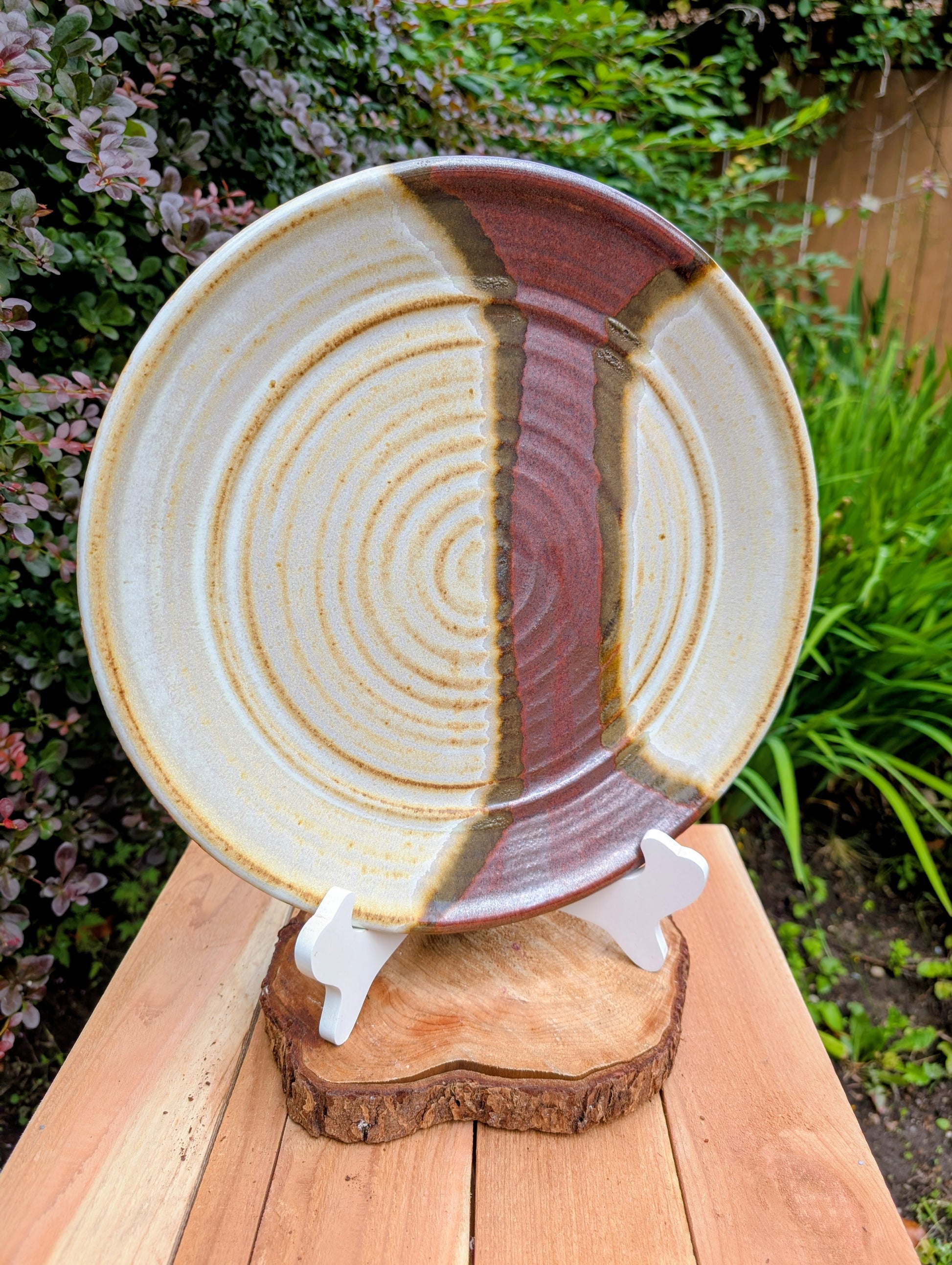 A striking hand-thrown studio pottery platter featuring an expressive cream, maroon, and olive glaze composition with visible throwing rings and an incised artist signature. Beautiful as both a decorative wall display and a functional serving piece, it embodies the warmth and craftsmanship of contemporary American studio ceramics.
