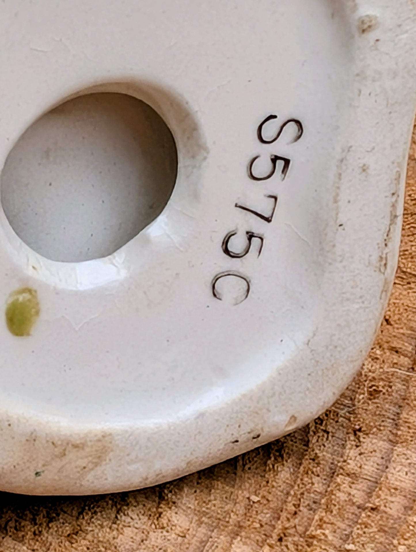 Close-up of the base of a white ceramic vessel, showing a circular hole and the printed black code 'S575C' on a rustic wood surface.