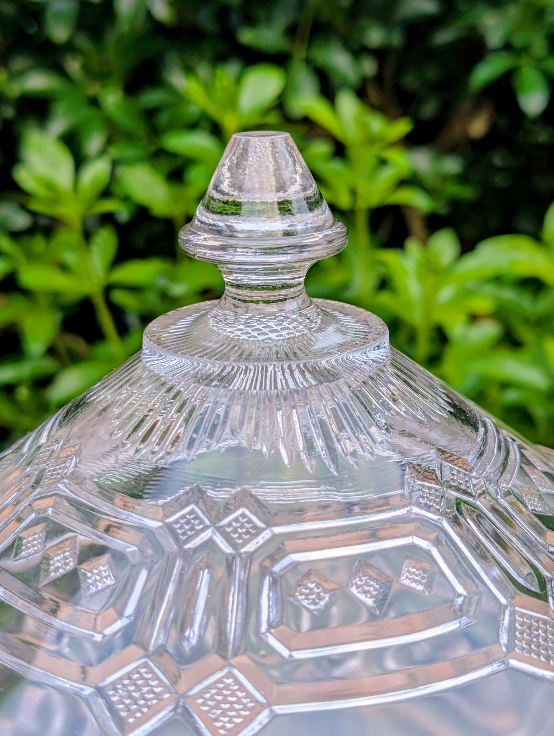 An elegant early 20th-century pressed glass covered compote featuring bold geometric Art Deco styling and a striking pedestal silhouette. Although the pedestal has visible stress cracks, the original lid, impressive scale, and architectural design make it a beautiful decorative display piece.