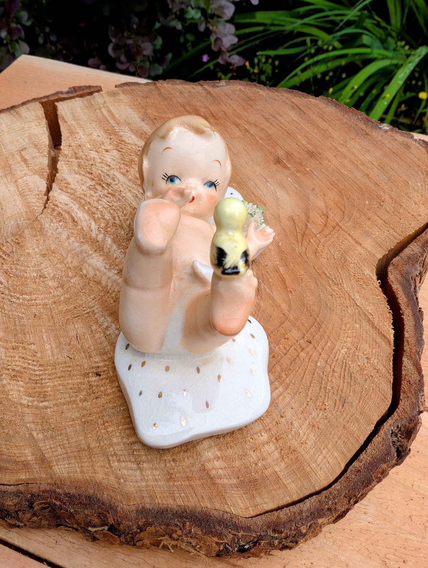 A vintage ceramic figurine of a baby holding a yellow-green acorn, displayed on a rustic wood slice with green foliage in the background.