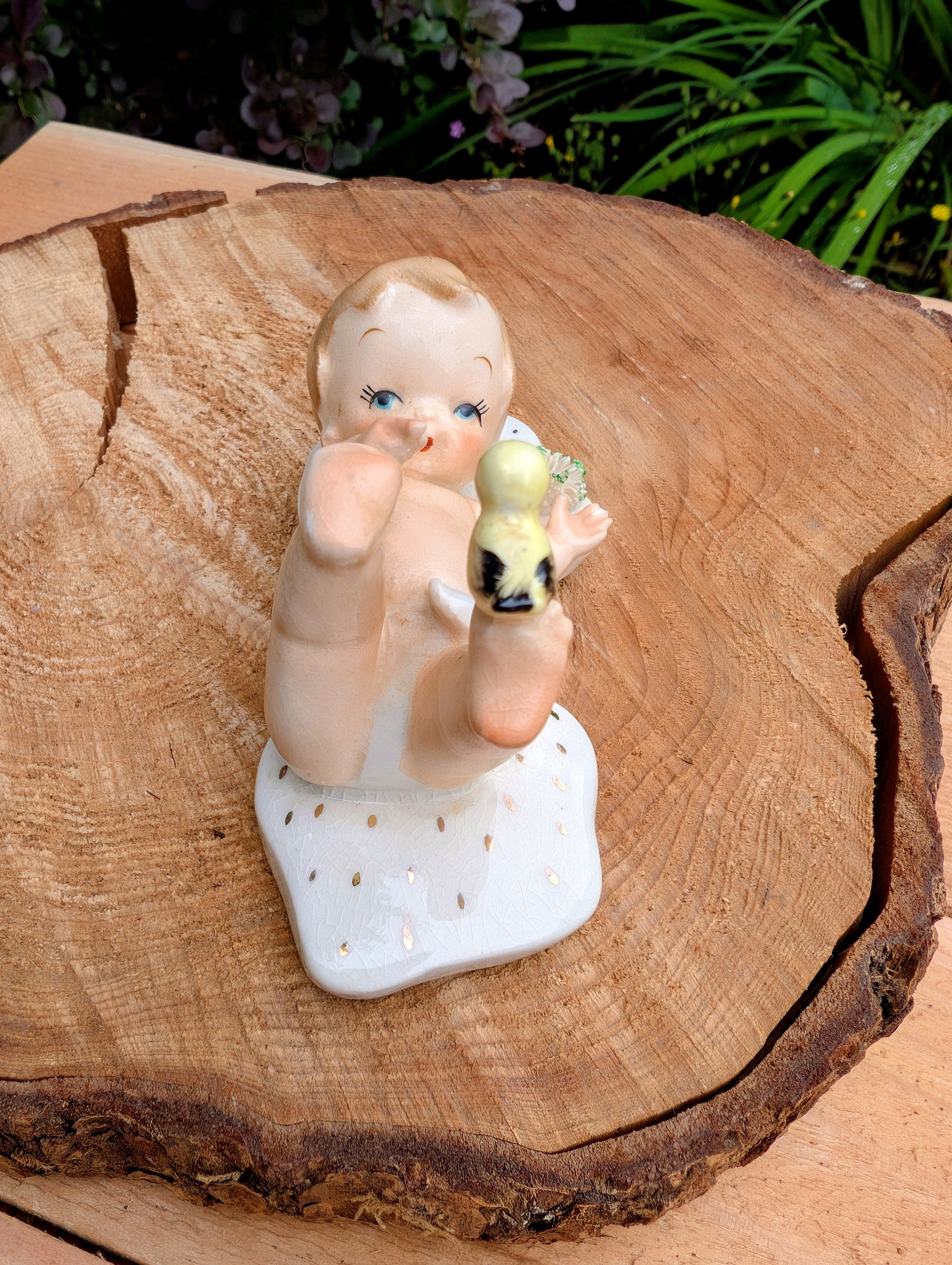 A vintage ceramic figurine of a baby holding a yellow-green acorn, displayed on a rustic wood slice with green foliage in the background.