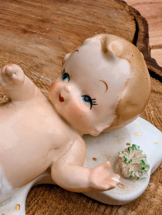 Vintage-style baby figurine lying on its back on a white cradle, placed on a rustic wood slice background. The figurine has a pale peach face and torso, light brown hair, blue eyes, and a small green-and-white flower accent.