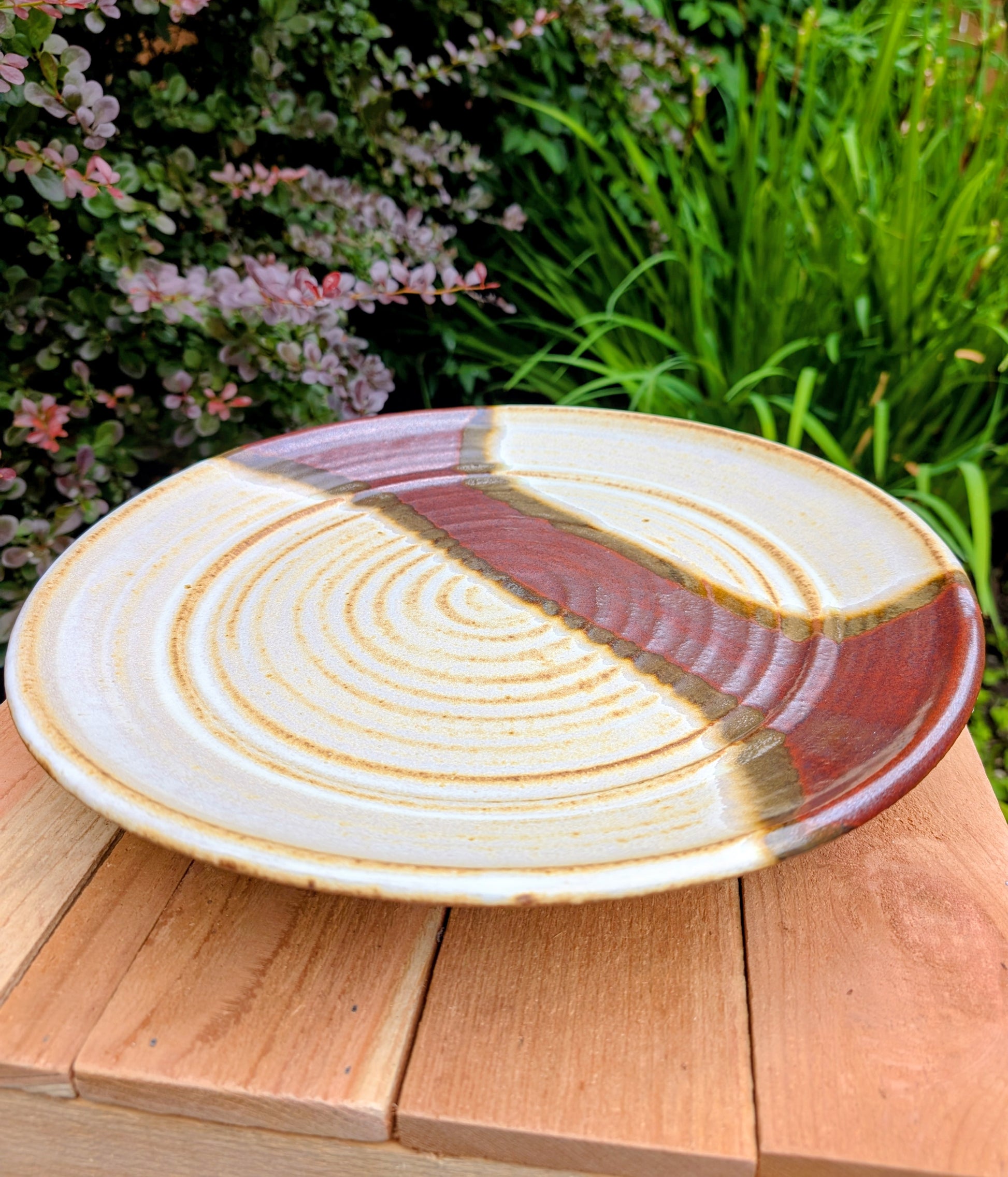 A striking hand-thrown studio pottery platter featuring an expressive cream, maroon, and olive glaze composition with visible throwing rings and an incised artist signature. Beautiful as both a decorative wall display and a functional serving piece, it embodies the warmth and craftsmanship of contemporary American studio ceramics.