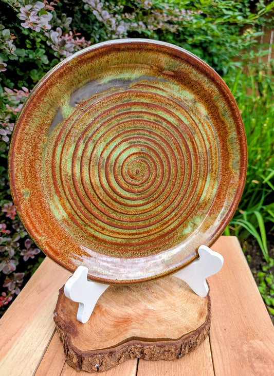 A beautifully hand-thrown studio pottery charger featuring an impressed spiral design and richly variegated moss green and rust glazes. This substantial signed stoneware piece brings timeless craftsmanship and Pacific Northwest-inspired warmth to shelves, tables, or wall displays.