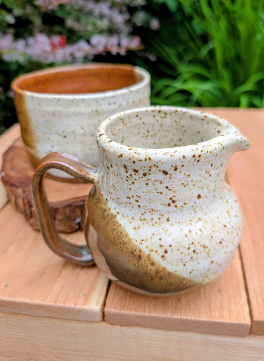 Bring handcrafted warmth to your table with this signed studio pottery creamer and sugar bowl set. Finished in a beautiful speckled cream glaze with flowing olive-brown accents, this artisan stoneware pair blends rustic craftsmanship with timeless everyday function.
