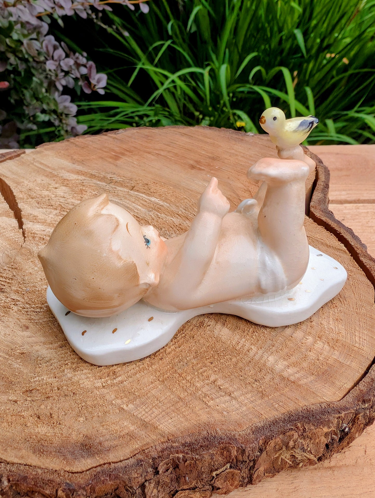 A ceramic figurine of a baby lying on its back on a white base, with a small yellow bird perched on the baby's foot, displayed on a rustic wood slice with green foliage in the background.