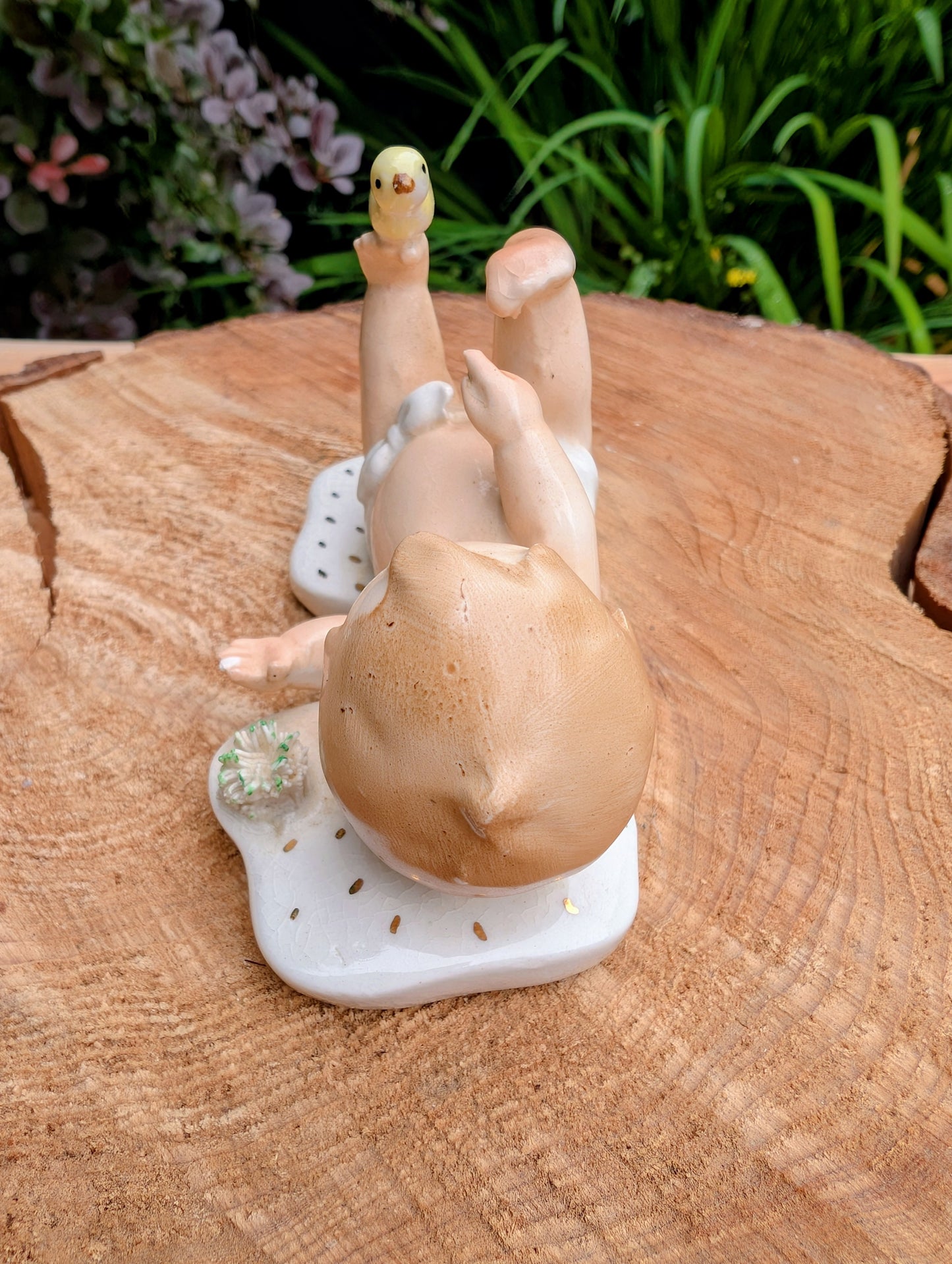 A small decorative figurine of a reclining baby with a yellow bird on its foot, displayed on a wooden surface with green foliage in the background.