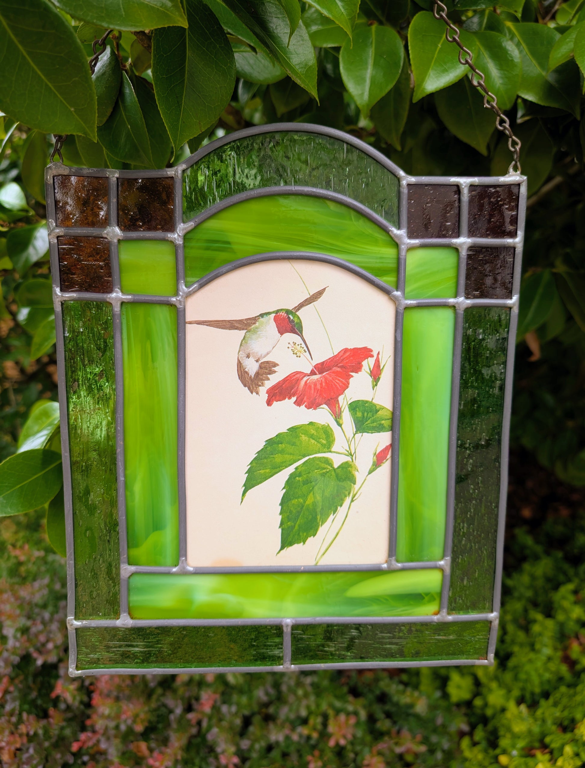 Bring the beauty of a hummingbird in bloom into your home with this handcrafted stained glass hanging panel. Featuring vibrant textured art glass surrounding a detailed hummingbird and hibiscus centerpiece, it catches natural light beautifully and adds year-round color to any window, sunroom, or garden-inspired space.